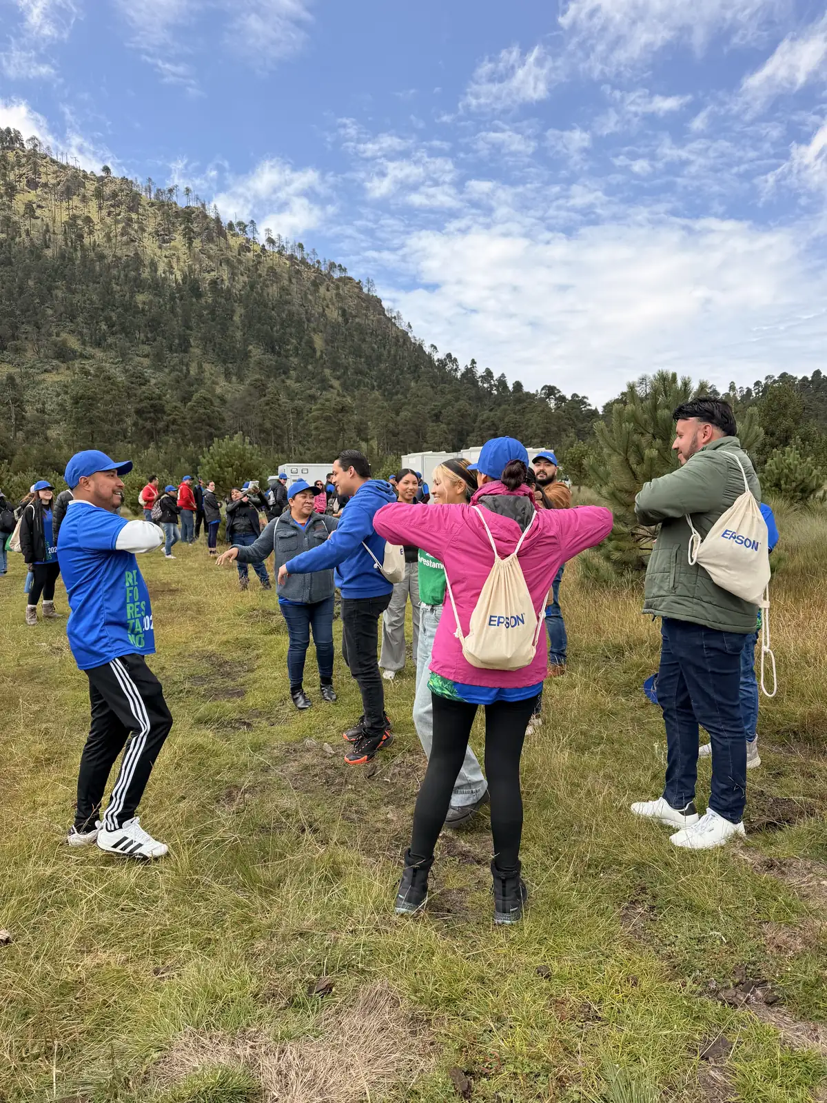 Voluntarios con gorras azules de Epson estiran los brazos en círculo sobre el pastizal, con el cerro boscoso al fondo