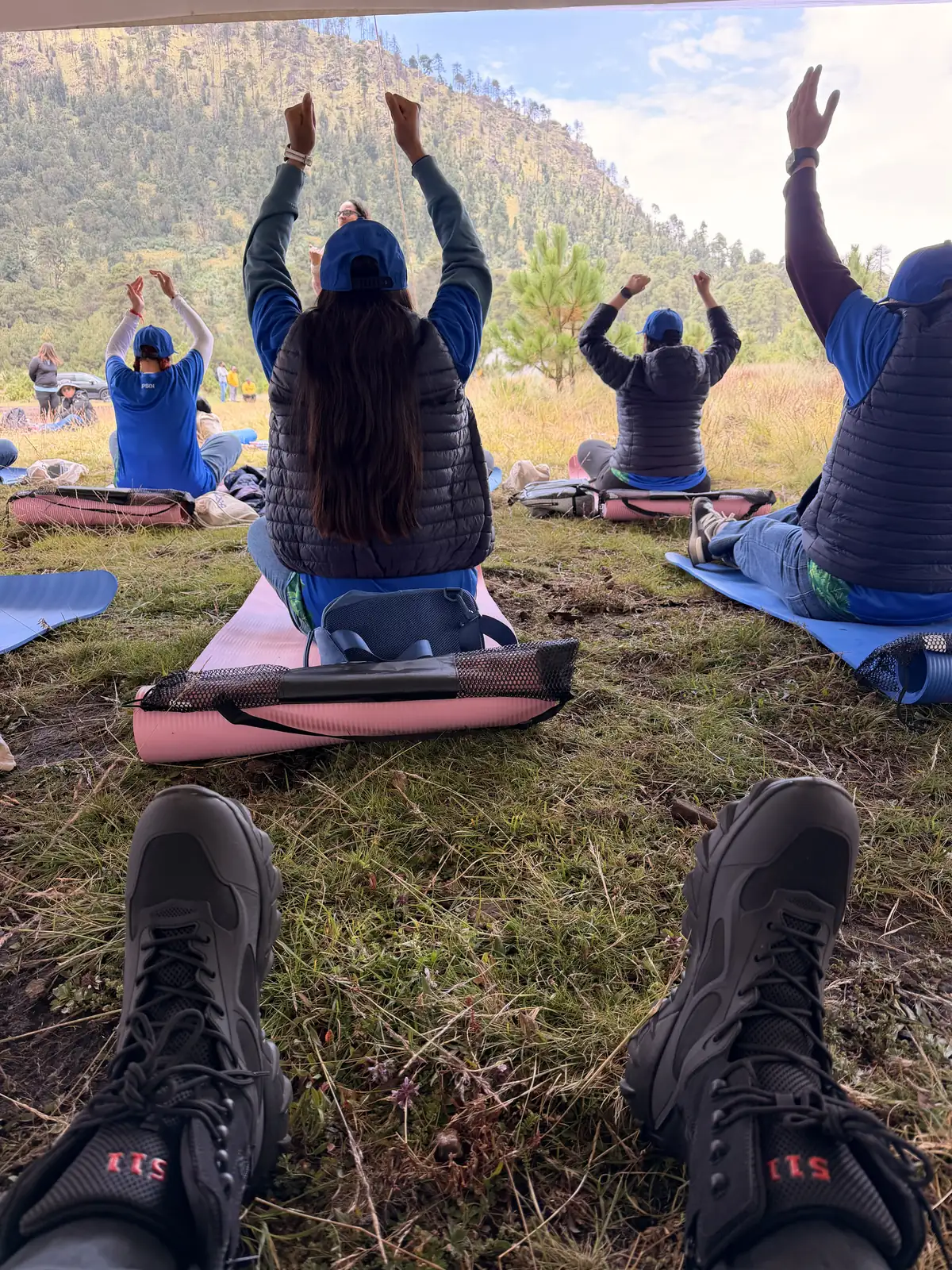 Voluntarios sentados sobre tapetes en el pasto con los brazos en alto durante la práctica de respiración guiada, de espaldas al bosque