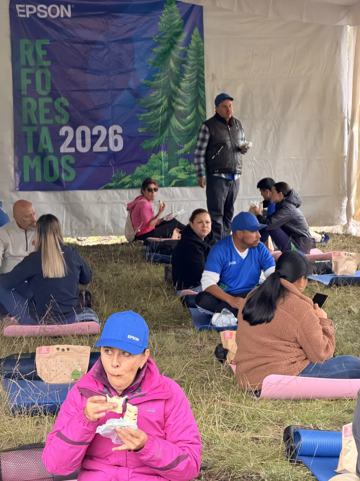 Voluntarios descansan sentados en tapetes bajo la carpa con la lona de Epson «Reforestamos 2026», comiendo después de la jornada
