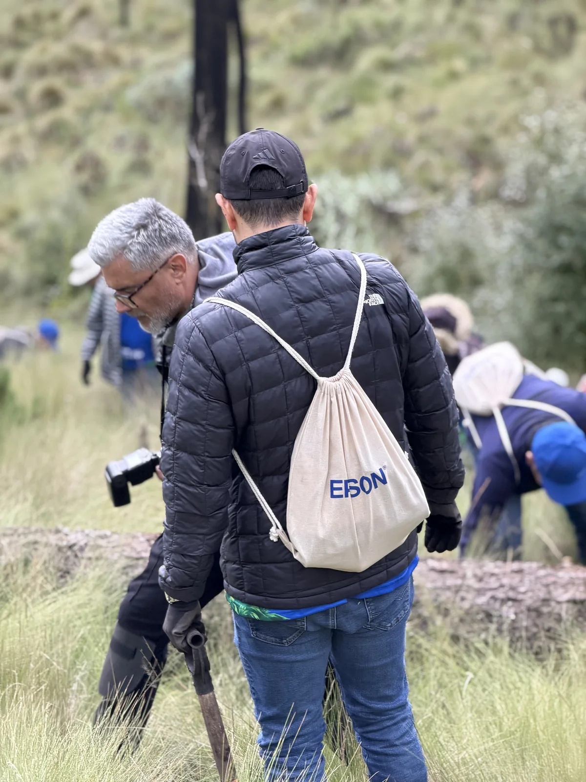 Integrante del equipo visto de espaldas, con un morral de tela con el logo Epson, caminando entre pinos y pastizal en la bajada