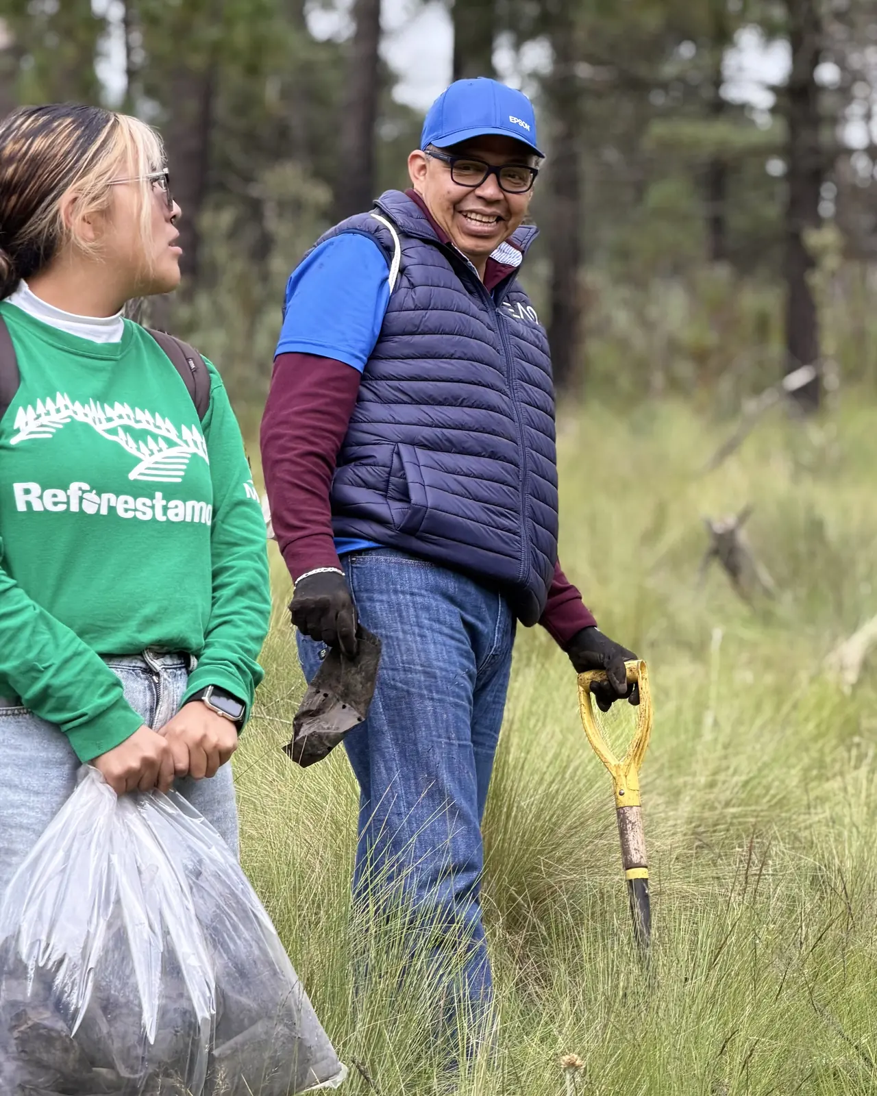 Dos integrantes del equipo, uno con bolsa de basura y otro con una herramienta, caminando entre el pastizal recogiendo desechos