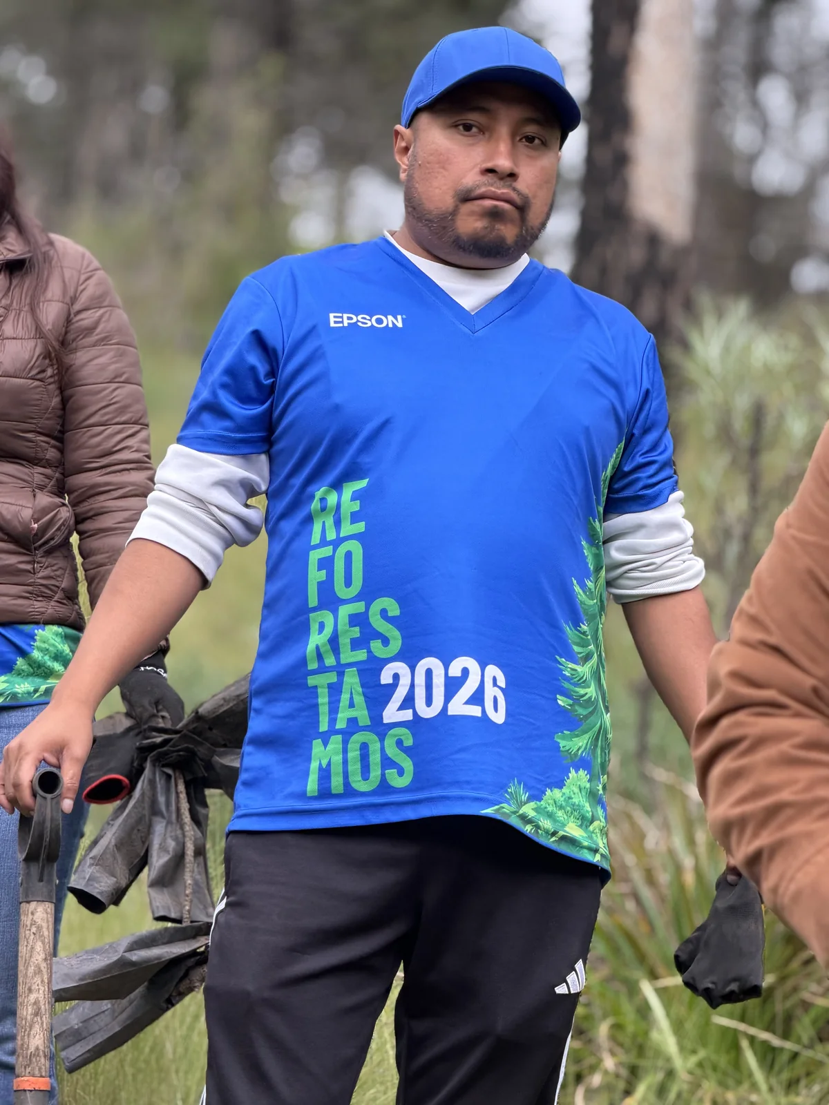 Integrante del equipo de espaldas con el jersey oficial «Reforestamos 2026», cargando una bolsa entre el pastizal